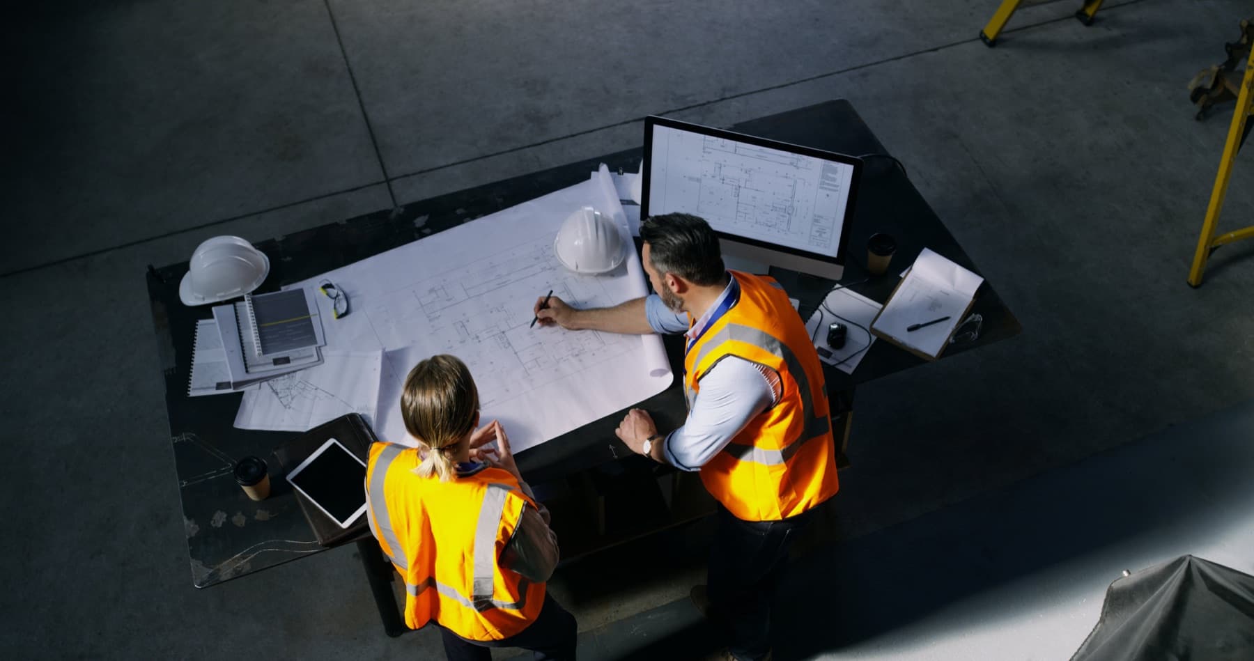 Two skilled workers in high-visibility vests review plans at a workstation.
